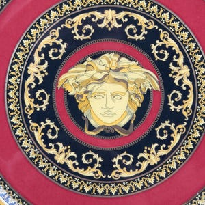 Op de afbeelding: Een decoratieve schotel met een rode kern en een zwarte rand met gouden krullen. Het midden van de schotel toont een Medusa-hoofd in gele en cr&egrave;metinten, een klassiek designelement.