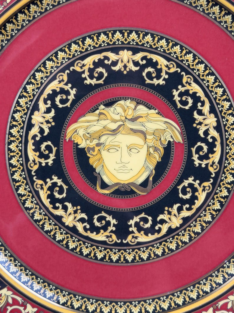 Op de afbeelding: Een decoratieve schotel met een rode kern en een zwarte rand met gouden krullen. Het midden van de schotel toont een Medusa-hoofd in gele en cr&egrave;metinten, een klassiek designelement.