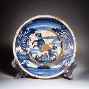 May include: An antique ceramic plate with a hand-painted design in blue and white. The plate features a central figure and floral motifs around the rim. The plate has a brown rim and is displayed on a clear stand.