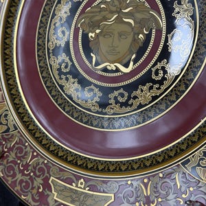 Op de afbeelding: Decoratieve plaat met een Medusa-hoofdontwerp in het midden. De plaat heeft een zwart, bordeauxrood en goud kleurenschema met sierlijke details. De plaat heeft een glanzende afwerking en is waarschijnlijk gemaakt van keramiek of porselein.
