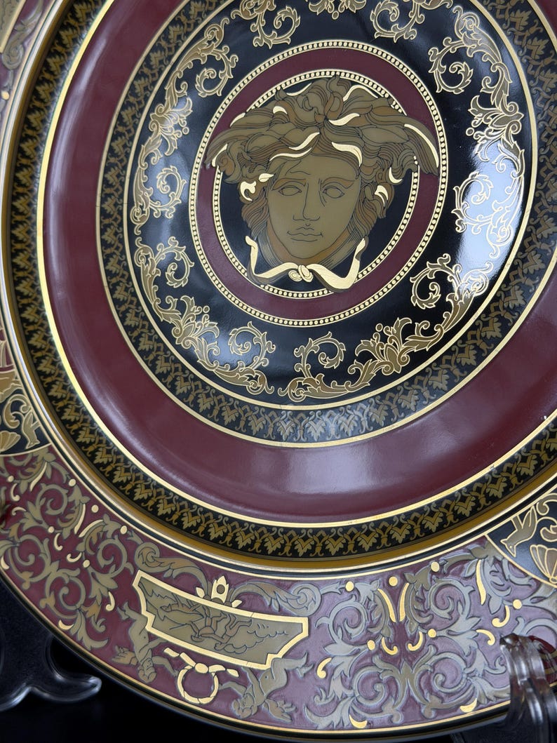 Op de afbeelding: Decoratieve plaat met een Medusa-hoofdontwerp in het midden. De plaat heeft een zwart, bordeauxrood en goud kleurenschema met sierlijke details. De plaat heeft een glanzende afwerking en is waarschijnlijk gemaakt van keramiek of porselein.