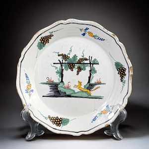 Peut inclure: Assiette en céramique blanche avec un bord festonné et un motif peint à la main. L'assiette représente un renard jaune, des vignes vertes et des raisins noirs. Le bord est souligné en noir et décoré d'accents floraux. L'assiette est présentée sur un support transparent.