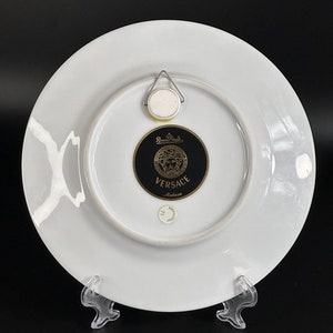 Puede incluir: Plato decorativo de porcelana blanca con el logotipo Versace Medusa en negro y dorado. El plato tiene un colgador de metal en la parte posterior. El plato est&aacute; sobre un soporte de pl&aacute;stico transparente.