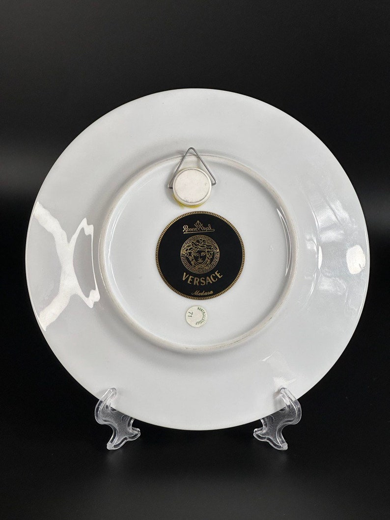 Puede incluir: Plato decorativo de porcelana blanca con el logotipo Versace Medusa en negro y dorado. El plato tiene un colgador de metal en la parte posterior. El plato est&aacute; sobre un soporte de pl&aacute;stico transparente.