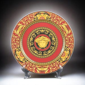 Op de afbeelding: Decoratieve plaat met een rood en goud ontwerp. De plaat heeft een centraal Medusa-hoofd embleem, omgeven door ingewikkelde patronen en een zwarte rand. De plaat wordt op een transparante standaard gepresenteerd.