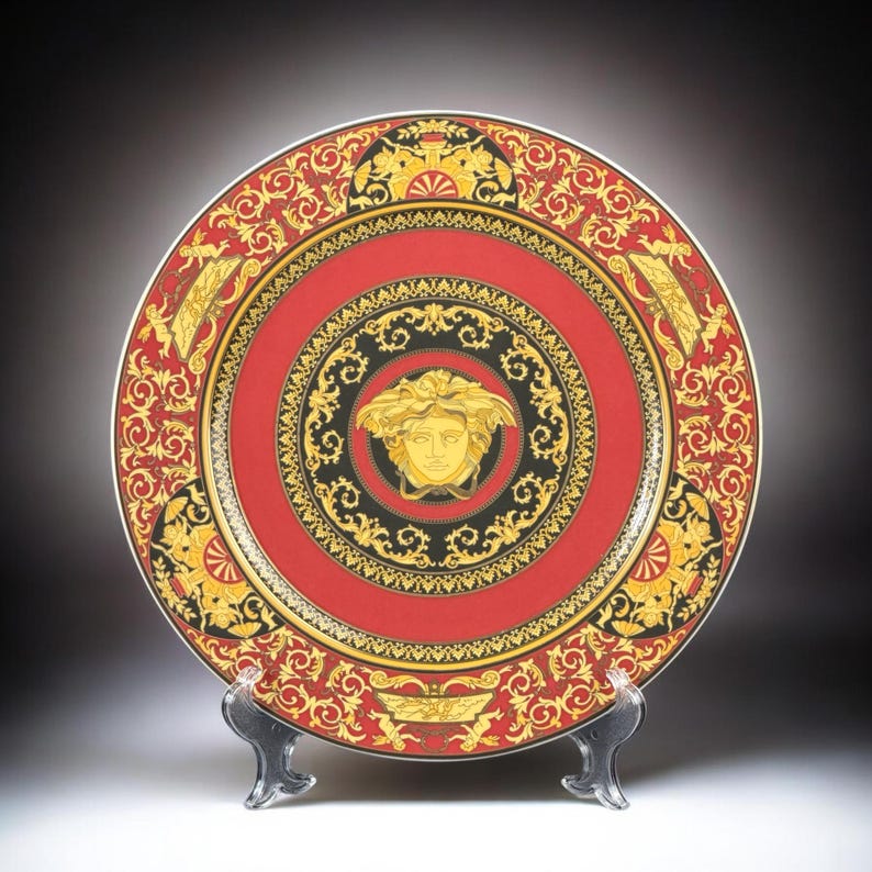 Op de afbeelding: Decoratieve plaat met een rood en goud ontwerp. De plaat heeft een centraal Medusa-hoofd embleem, omgeven door ingewikkelde patronen en een zwarte rand. De plaat wordt op een transparante standaard gepresenteerd.