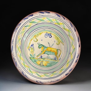 May include: A round ceramic bowl featuring a hand-painted bull in the center, colored in yellow and green. The bowl is decorated with concentric blue and green rings, leafy patterns, and a speckled blue rim on a cream-colored glazed surface.