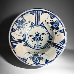 May include: A white ceramic plate with a blue floral design. The plate features a central floral motif surrounded by radiating lines and additional floral accents. The rim has a scalloped edge with blue detailing. The plate is likely vintage or antique.