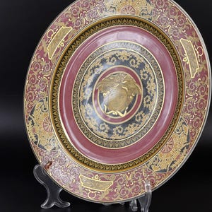 Op de afbeelding: Decoratieve plaat met een bordeauxrood en goudkleurig ontwerp. De plaat heeft een centraal medaillon met een gouden gezicht, omgeven door concentrische cirkels en sierlijke patronen. De rand heeft een gouden en bordeauxrood bloemmotief.