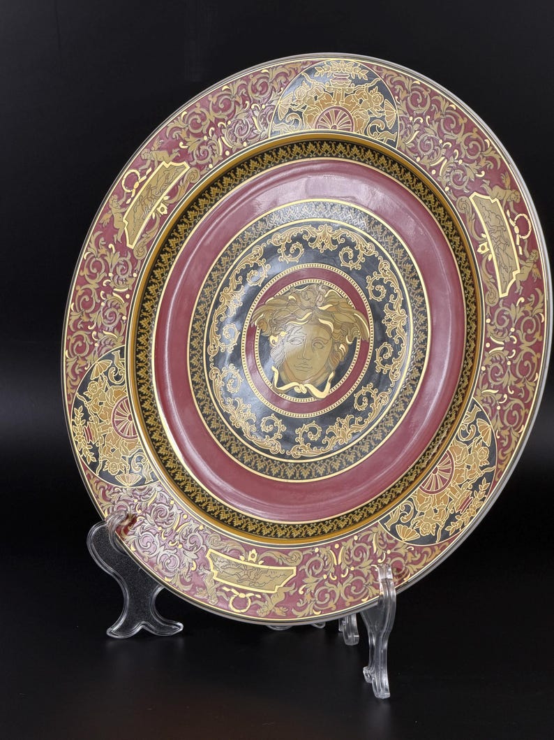 Op de afbeelding: Decoratieve plaat met een bordeauxrood en goudkleurig ontwerp. De plaat heeft een centraal medaillon met een gouden gezicht, omgeven door concentrische cirkels en sierlijke patronen. De rand heeft een gouden en bordeauxrood bloemmotief.