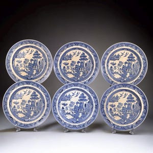 May include: A set of six blue and white porcelain plates, each with a Willow pattern design. The plates depict a landscape scene with a pagoda, trees, and birds, framed by a patterned border. The plates are presented on clear stands.