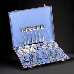 May include: A collection of silver-coloured cutlery, including spoons and forks, is arranged inside an open, rectangular case lined with blue fabric. The silverware features decorative detailing. The case is brown with a gold clasp.
