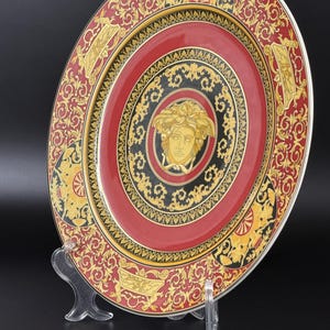 Op de afbeelding: Decoratieve plaat met een rood, zwart en goud ontwerp. De plaat heeft een centrale Medusa-kop omringd door sierlijke patronen en een gouden rand. De plaat wordt gepresenteerd op een transparante standaard tegen een zwarte achtergrond.