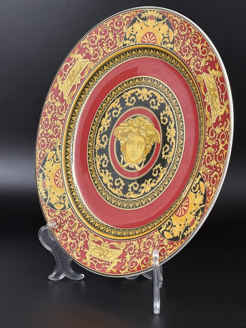 Op de afbeelding: Decoratieve plaat met een rood, zwart en goud ontwerp. De plaat heeft een centrale Medusa-kop omringd door sierlijke patronen en een gouden rand. De plaat wordt gepresenteerd op een transparante standaard tegen een zwarte achtergrond.