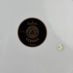 Op de afbeelding: Een witte porseleinen plaat met een zwart en goud Versace Medusa-logo. Het logo is gecentreerd en toont de naam Versace en het Medusa-hoofd. Een kleine, ronde sticker met het nummer 70 is ook zichtbaar.