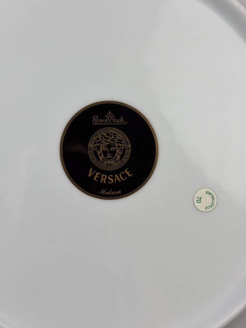 Op de afbeelding: Een witte porseleinen plaat met een zwart en goud Versace Medusa-logo. Het logo is gecentreerd en toont de naam Versace en het Medusa-hoofd. Een kleine, ronde sticker met het nummer 70 is ook zichtbaar.