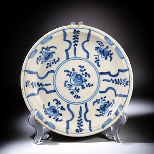 May include: Antique ceramic plate with a white base and blue floral patterns. The plate features a central floral design surrounded by a circular border and repeating floral motifs. The rim has a wavy blue design. The plate is displayed on a clear stand.