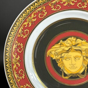 Puede incluir: Primer plano de un plato decorativo con un dise&ntilde;o negro, rojo, blanco y dorado. El plato presenta una cabeza de Medusa dorada en el centro, rodeada por un borde rojo y negro. El borde exterior tiene un patr&oacute;n dorado y rojo.