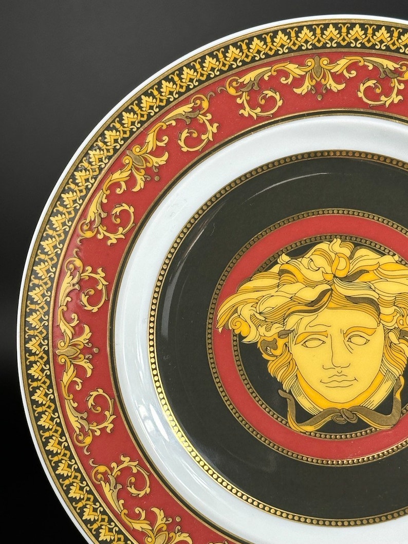 Puede incluir: Primer plano de un plato decorativo con un dise&ntilde;o negro, rojo, blanco y dorado. El plato presenta una cabeza de Medusa dorada en el centro, rodeada por un borde rojo y negro. El borde exterior tiene un patr&oacute;n dorado y rojo.