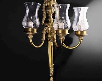 Può includere: Applique da parete in ottone dorato con tre paralumi in vetro trasparente. L'applique presenta un design dettagliato con bracci curvi e dettagli decorativi. Ogni paralume in vetro ha un motivo inciso. Sfondo scuro.