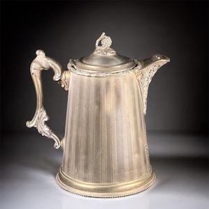 May include: Silver-colored antique coffee pot with a lid and a decorative handle. The pot has vertical striped detailing and ornate accents on the handle and spout. The lid is topped with a small, decorative finial.