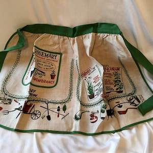 May include: A vintage apron with a beige background and green trim. The apron features illustrations of herbs like rosemary, chives, and marjoram, along with gardening tools. Text includes "Rosemary," "Chives," and "Sweet Marjoram."