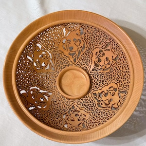 Może przedstawiać: Okrągły, drewniany talerz w jasnobrązowym kolorze. Talerz posiada szczegółowy, wycinany laserowo wzór przedstawiający wilcze twarze. Środek ma małe, okrągłe wgłębienie. Zewnętrzna krawędź jest podniesiona.