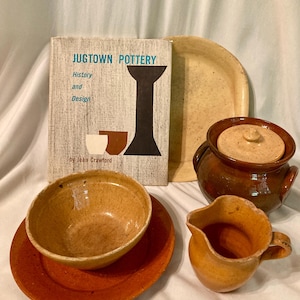 May include: A collection of brown and tan pottery pieces, including a bowl, a pitcher, a covered jar, and a plate. A book titled "Jugtown Pottery: History and Design" by Jean Crawford is also visible.