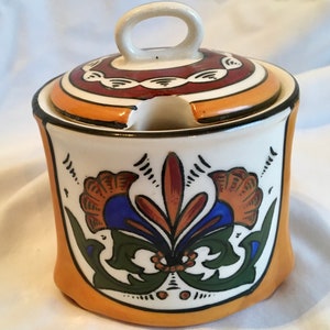 Może przedstawiać: Ceramiczna cukiernica z pokrywką, ozdobiona kwiatowym wzorem w odcieniach niebieskiego, zielonego, pomarańczowego i brązowego. Cukiernica ma białe tło z żółtym obramowaniem.