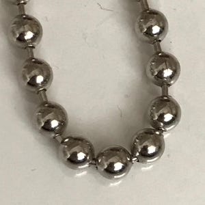 May include: Close-up of a shiny, silver ball chain. The chain is made up of many small, spherical metal beads connected by short, straight links. The beads reflect light, creating highlights and shadows.