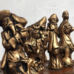 Può includere: Un set di sei pezzi degli scacchi dipinti in oro, con tema pirata. I pezzi sono disposti su una scacchiera di legno con intagli intricati.