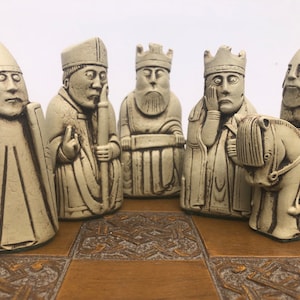 May include: A set of off-white chess pieces with detailed facial expressions and attire, including a king, queen, and bishop. The pieces are arranged on a wooden chessboard with a decorative pattern. The chess pieces are approximately 3 inches tall.