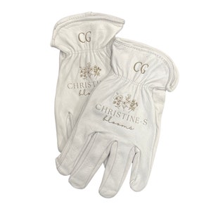 Puede incluir: Un par de guantes de trabajo de cuero blanco con el texto "CHRISTINE'S blooms" y "CG" bordado.