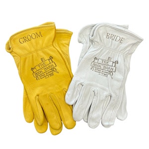 May include: A pair of leather work gloves, one yellow and one white, with the words "Groom" and "Bride" embroidered on the back. The gloves have a logo on the palm that says "The Book Barn" with an empty slogan.