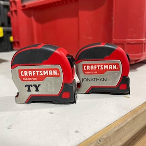 May include: Two red and black Craftsman tape measures with silver accents. The tape measure on the left has the name "TY" printed on it. The tape measure on the right has the name "JONATHAN" printed on it.