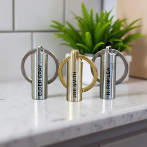 Könnte beinhalten: Drei personalisierte Schlüsselanhänger mit zylindrischen Metallbehältern. Die Schlüsselanhänger sind silber- und goldfarben und tragen die Namen "ELDER GRAY", "JOE SMITH" und "R. FINKLEA" vertikal aufgedruckt. Eine grüne Pflanze in einem weißen Topf befindet sich im Hintergrund.