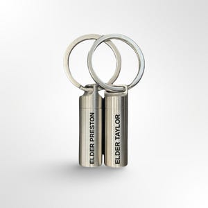 May include: Two silver-colored keychains with cylindrical pendants. Each pendant has a silver keyring attached. One keychain reads "ELDER PRESTON" and the other reads "ELDER TAYLOR" in black text. The background is white.