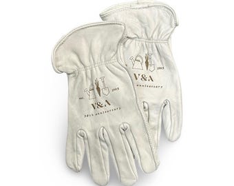 May include: A pair of off-white leather gardening gloves. The gloves have a brown logo that reads "V&A" with gardening tools and the text "est. 1995 30th anniversary". The gloves are designed for gardening and yard work.