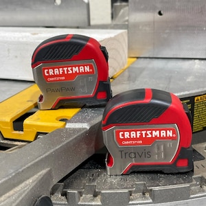 May include: Two red and black Craftsman tape measures with silver accents. The top tape measure has the name "PawPaw" and the bottom tape measure has the name "Travis".