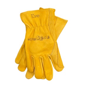 May include: A pair of yellow leather work gloves with the name "Eve" embroidered on the back of the left glove. The gloves feature a mountain range design on the back of the left glove.