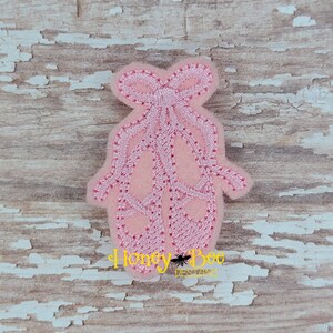 May include: Pink felt ballerina shoes with a pink ribbon bow. The shoes are embroidered with a stitch detail.