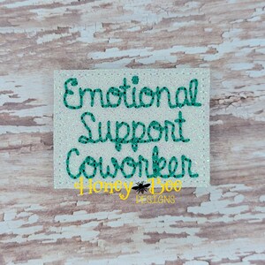 May include: A white glitter background with the text "Emotional Support Coworker" in teal green lettering. The text is in a cursive font. The text is on a white background with glitter.