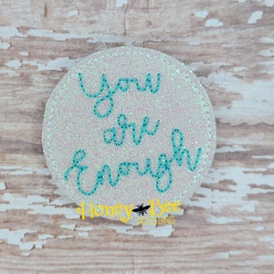 May include: White glitter circle with the text "you are enough" in turquoise blue cursive lettering. The text is centered on the circle. The circle is on a wood background.