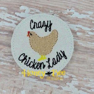 May include: A white circle with glitter, featuring a brown chicken with a red comb and yellow feet. The text "Crazy Chicken Lady" is written in black cursive font.