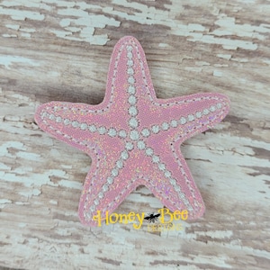 May include: A pink glittery starfish with white stitching and a white outline. The starfish is on a light brown wooden background. Honey Bee Designs is written in white text below the starfish.