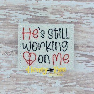 May include: White glitter background with red and black embroidered text that reads "He's still working on me" with a red heart with a black cross inside.