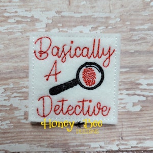 May include: White felt patch with red embroidered text that reads "Basically a Detective" with a black magnifying glass with a red fingerprint inside. The text "Honey Bee Designs" is embroidered in yellow at the bottom.