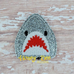 May include: A silver glitter shark face with a red open mouth and black eyes. The shark face is embroidered on a white fabric.
