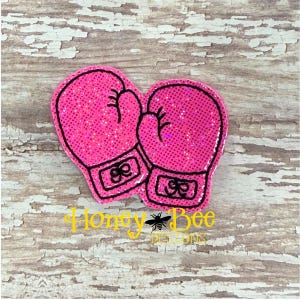 May include: A pair of bright pink glittery boxing gloves with black stitched outlines and detailing. The gloves are positioned side-by-side, featuring a textured surface with a sparkling finish and a small rectangular wrist strap area on each glove.