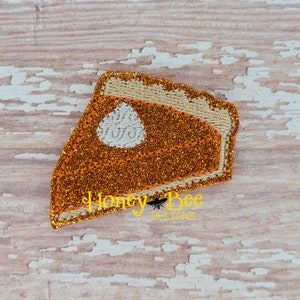 May include: A slice of pumpkin pie with a dollop of whipped cream, embroidered with glitter and a brown stitched outline. The pie is on a woodgrain background.